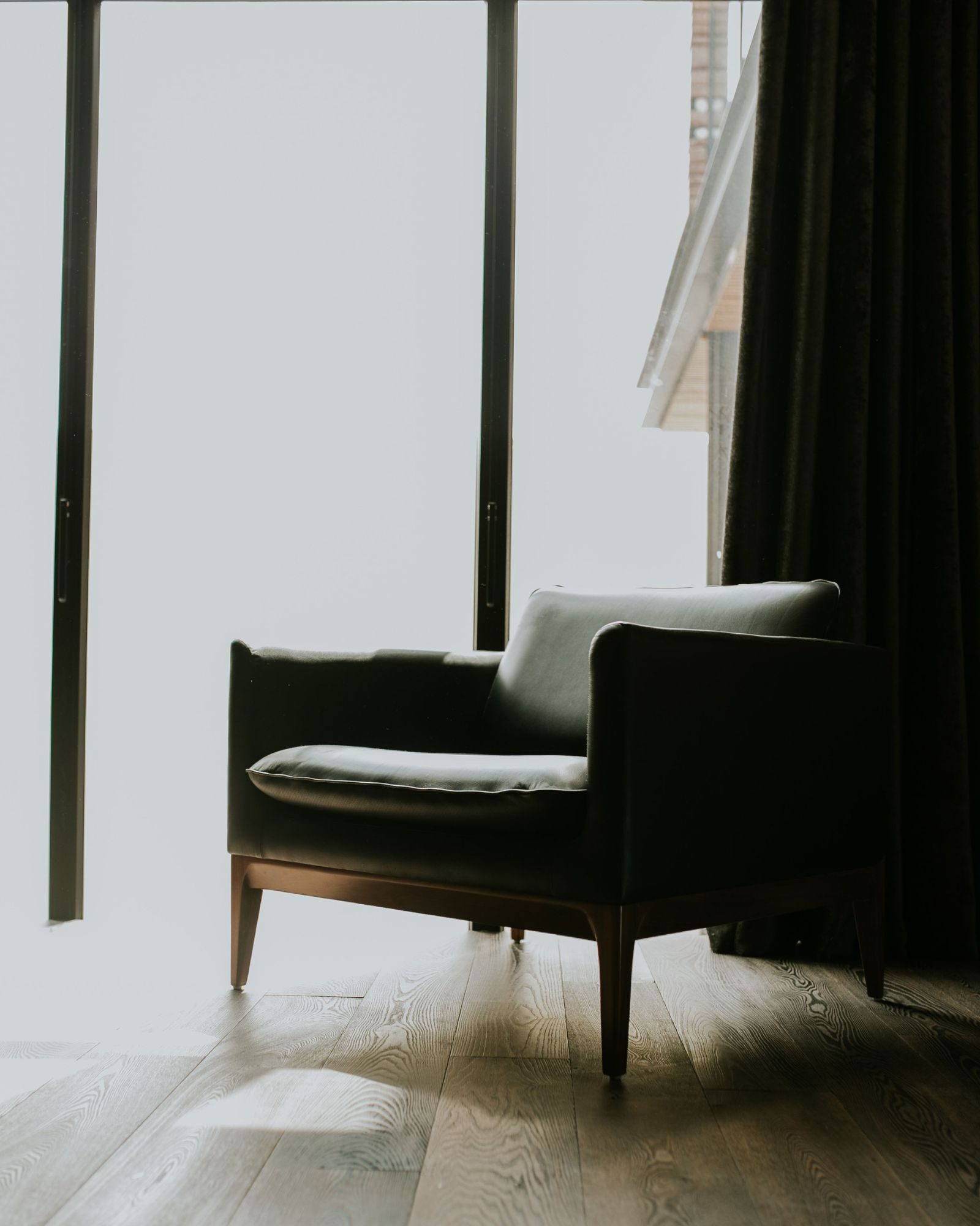 A single dark leather armchair by a tall bright window, a heavy curtain beside it